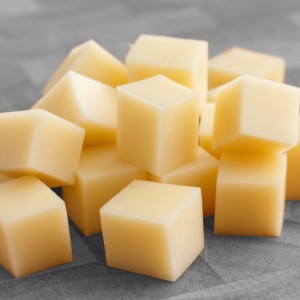 Cubes of yellow cheese stacked randomly on wood chopping board.