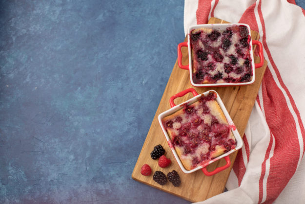Homemade Raspberry and Blueberry Cobblers in Red Cookware with Red and White Kitchen Towel against a Blue Background